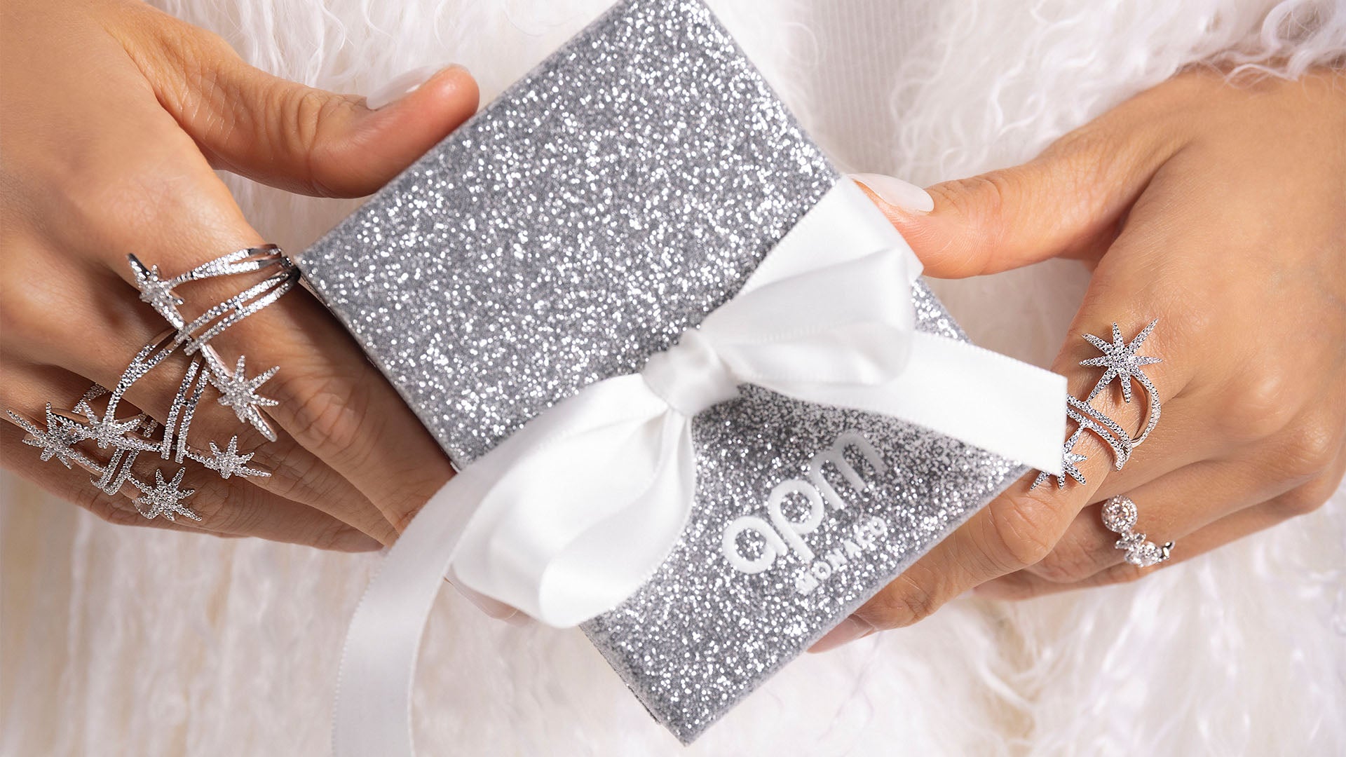 Silver glittery gift box with white ribbon held by hands, featuring the brand 'APM MONACO'