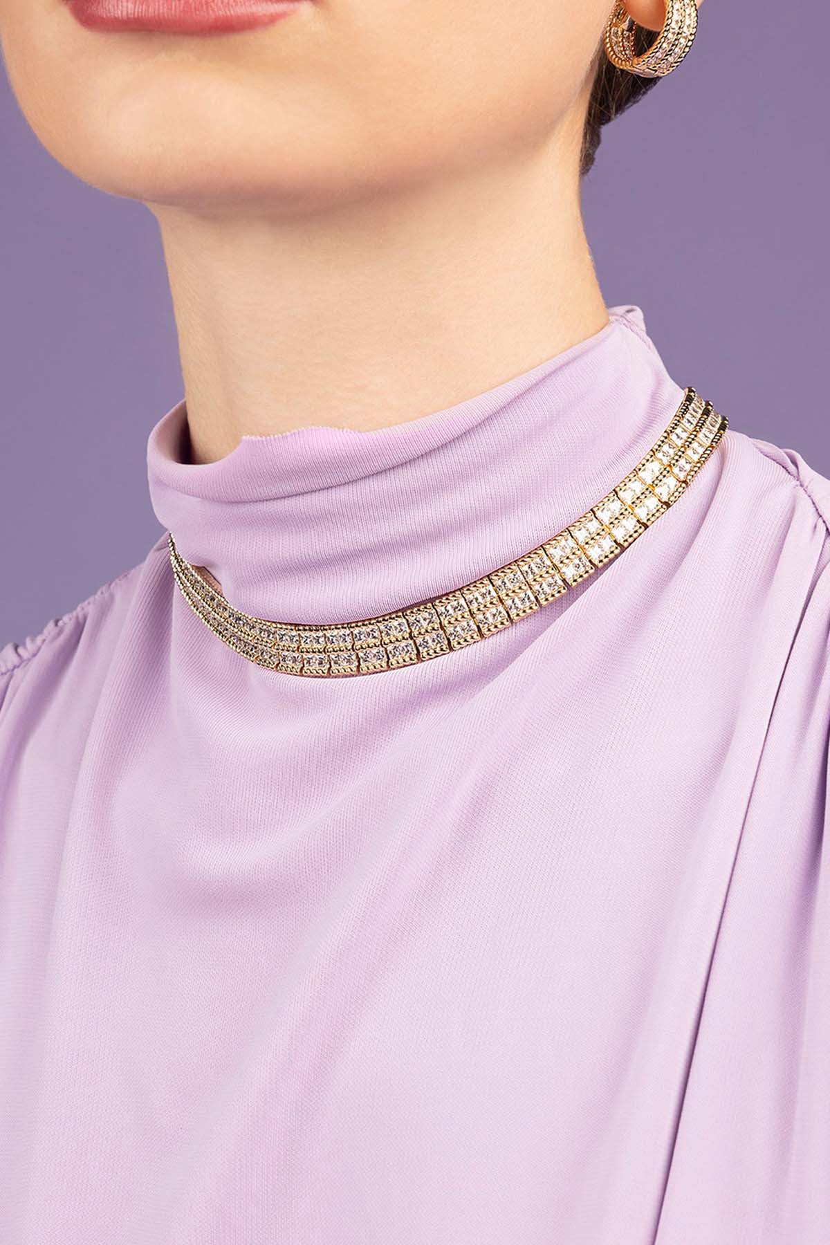 Close-up of a person wearing a lavender top with a decorative gold neckline against a purple background.