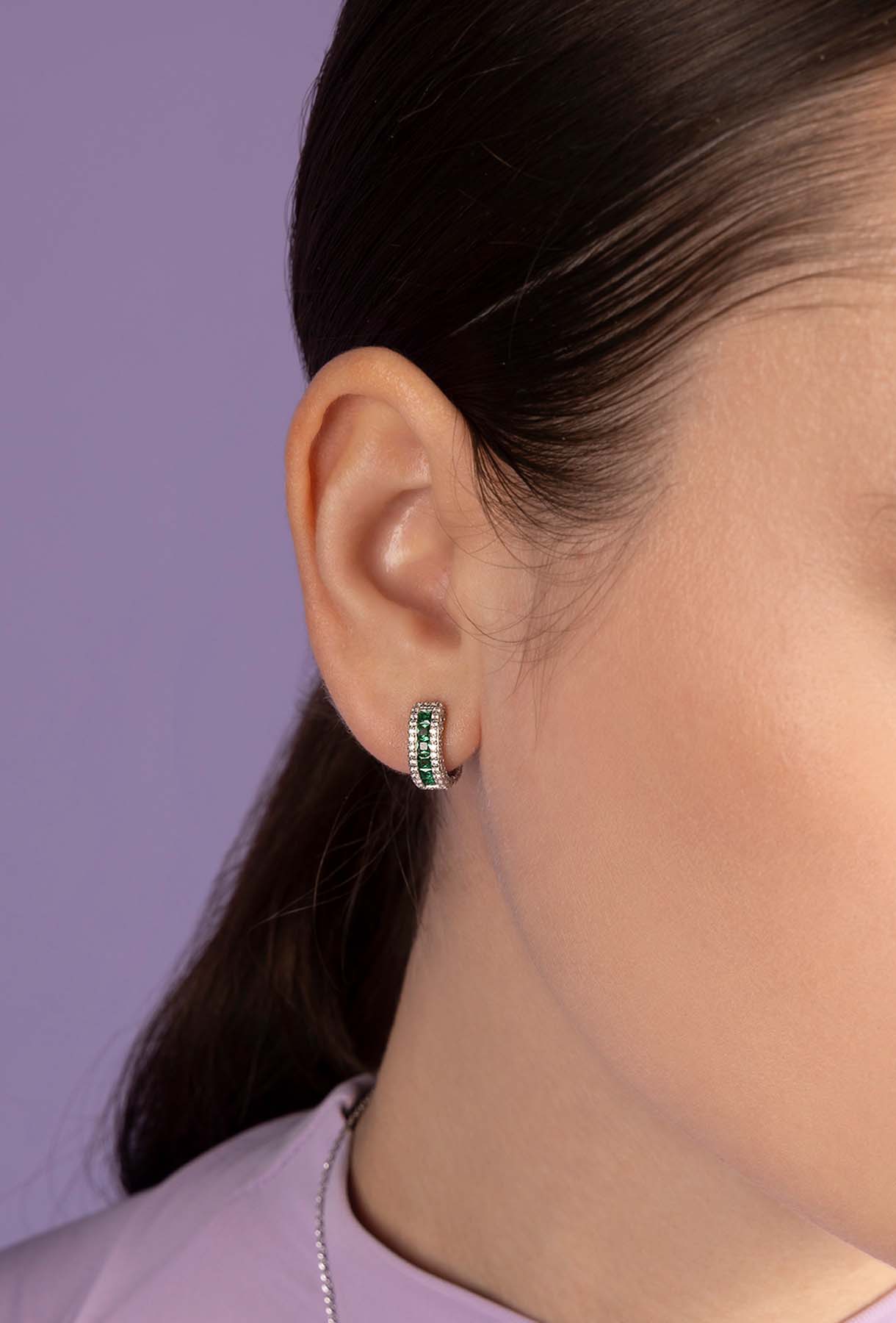 Close-up of an ear wearing APM MONACO green pave huggie earrings with a blurred purple background
