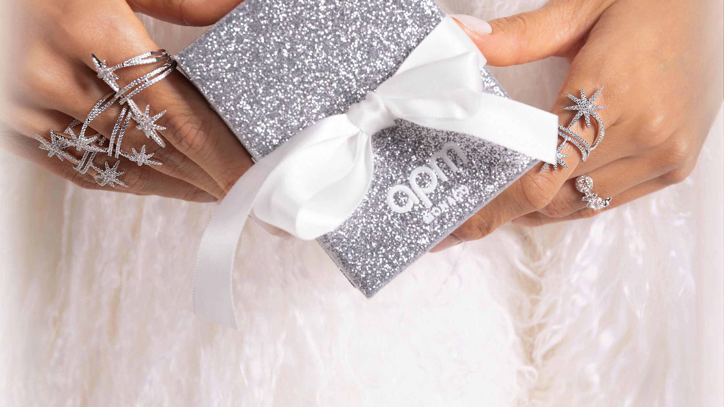 Silver APM MONACO glittery gift box with a white ribbon held by hands wearing star-shaped rings on a light background.