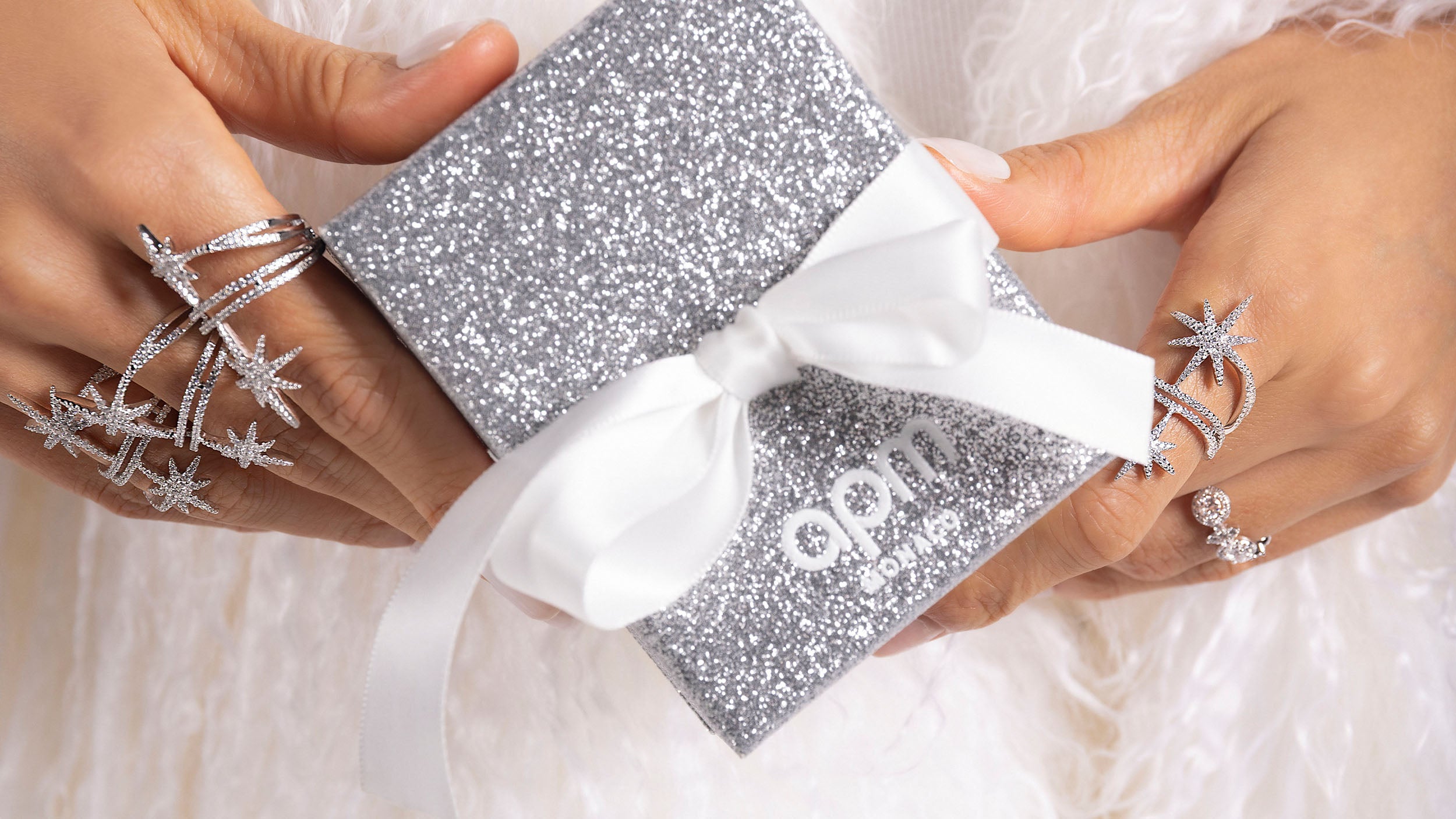 Silver APM MONACO glittery gift box with a white ribbon held by hands wearing star-shaped silver rings on a light background.