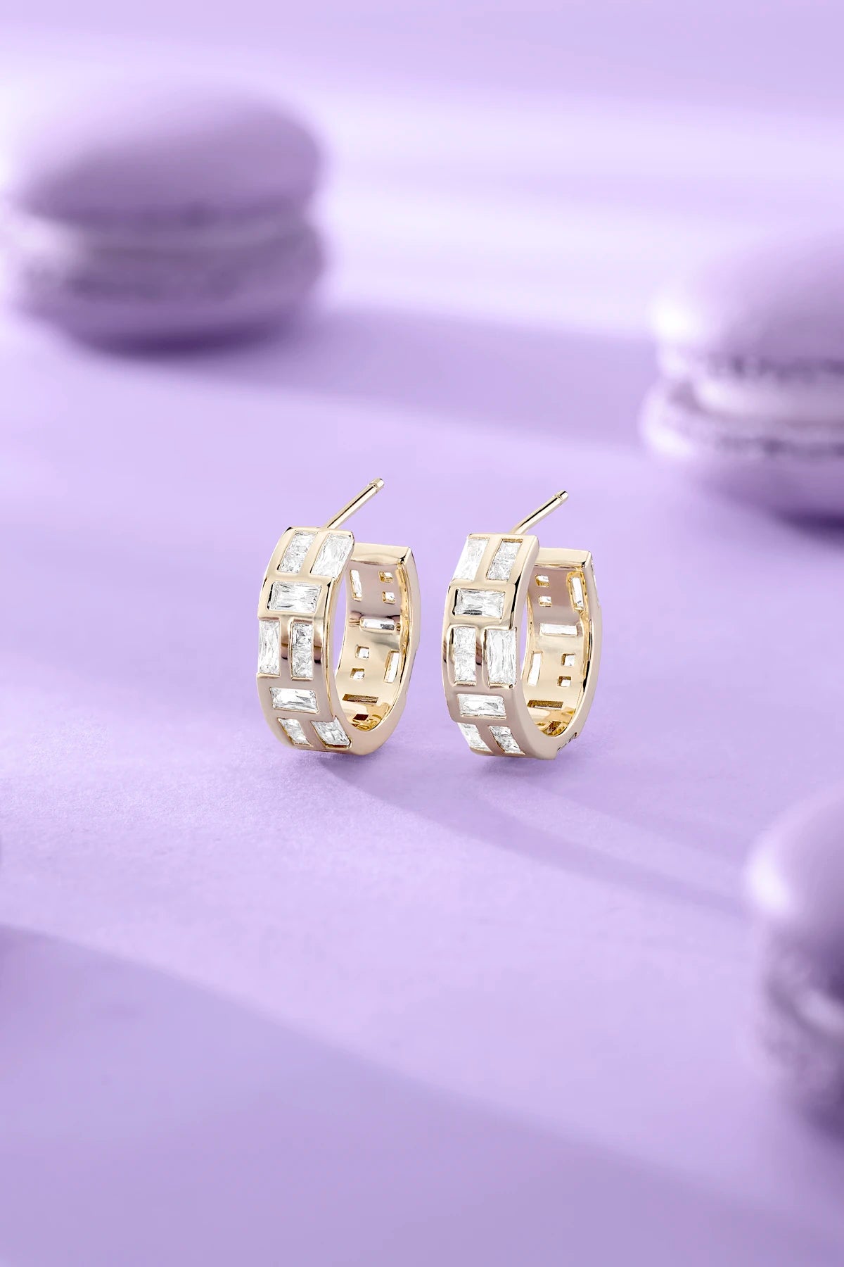 APM MONACO yellow gold hoop earrings on a light purple surface with blurred macarons in the background